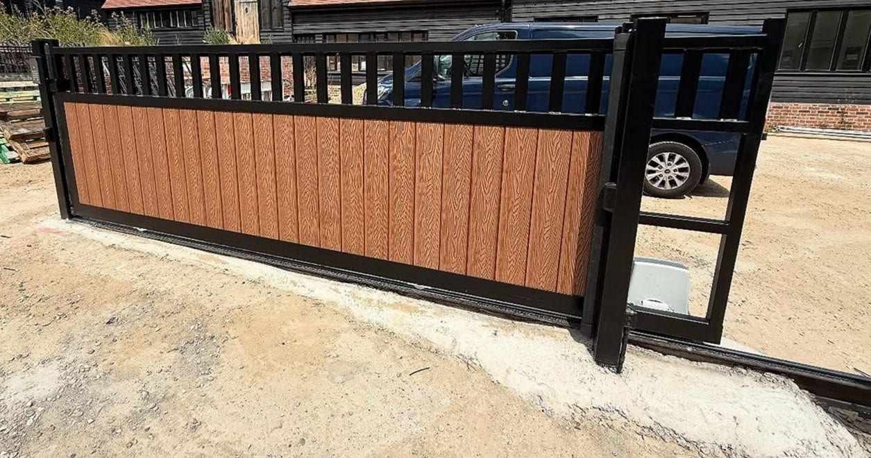 New sliding gate with wood-grain composite panels in a black steel frame, set across a freshly graded drive