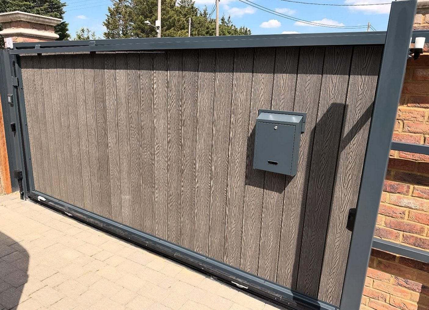 Close view of a sliding gate in dark wood-look boards with a mailbox fixed to the frame, beside a brick pier