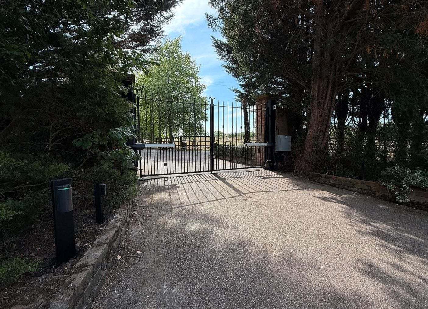 Black metal double swing gate at the end of a tree-lined driveway, low bollard lights along the drive