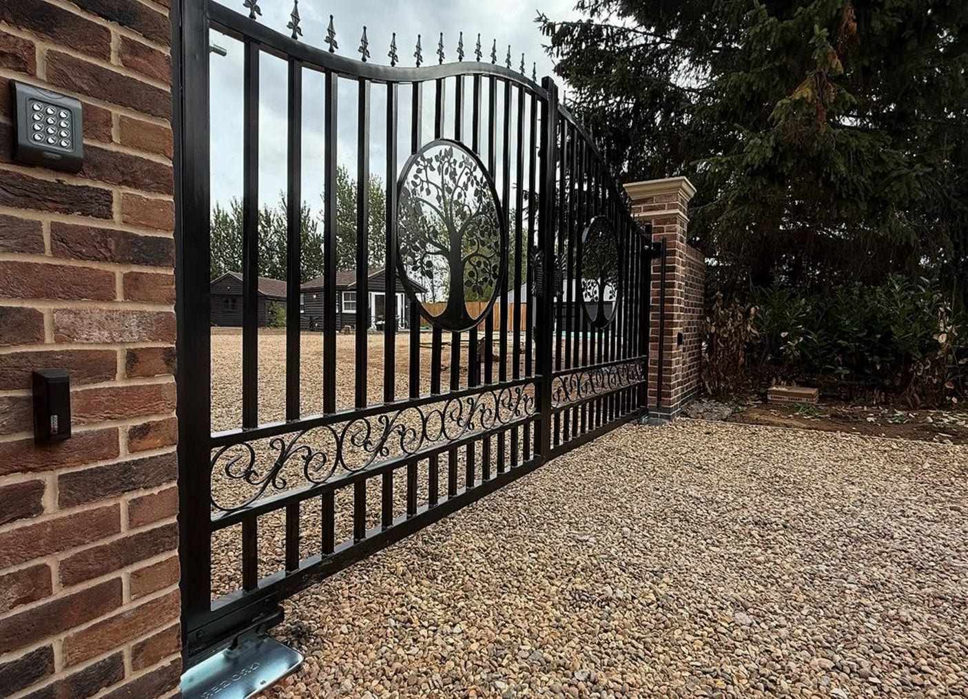Black steel sliding gate with a tree-of-life panel and scrollwork, a keypad and a push button mounted on the brick pier beside it