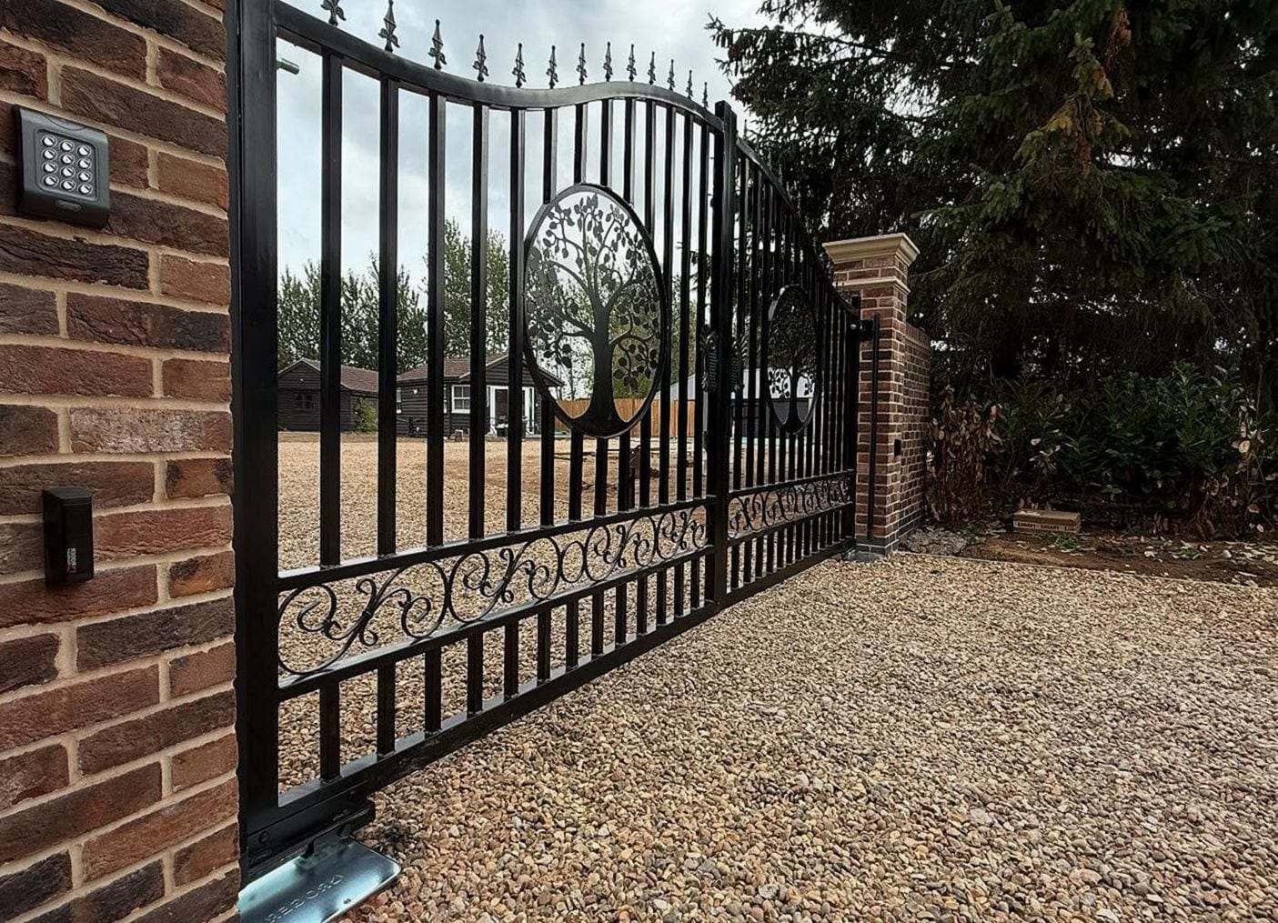 Black steel sliding gate with a tree-of-life panel and scrollwork, a keypad and a push button mounted on the brick pier beside it