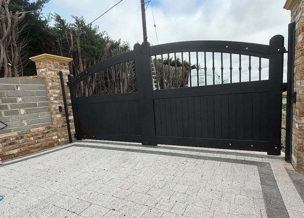 Black arched-top driveway gate between stone-faced piers, closed across new paving