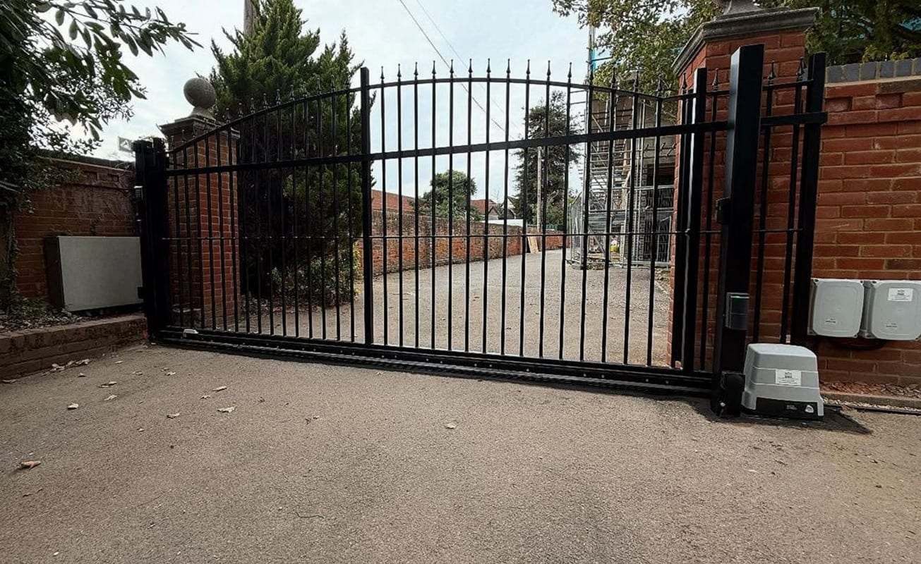Black sliding driveway gate with arched spear-top pickets between brick walls, the gate motor on its pad at the right