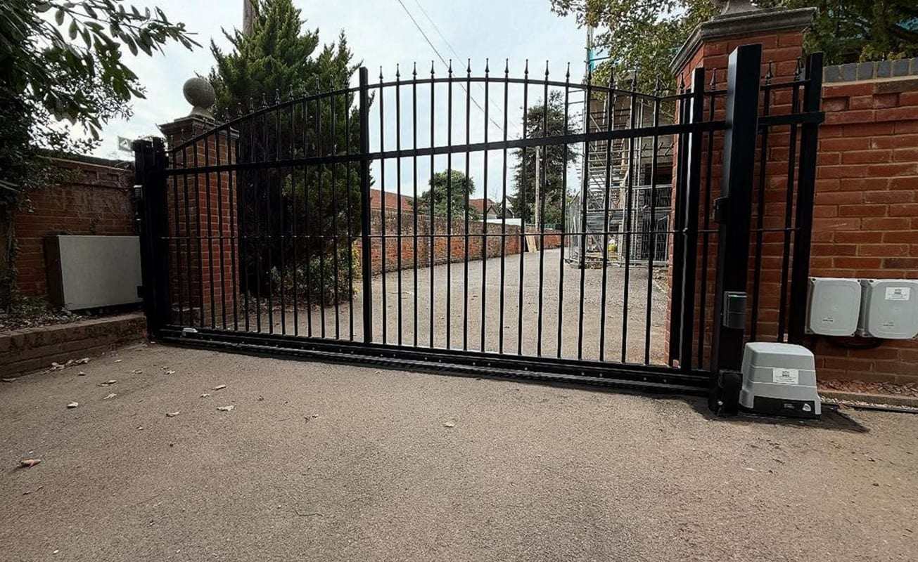 Black sliding driveway gate with arched spear-top pickets between brick walls, the gate motor on its pad at the right