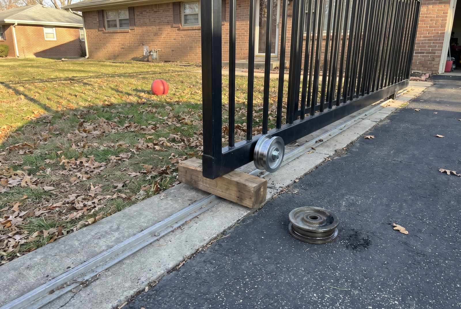 Sliding driveway gate propped on a wooden block during a roller repair, a worn wheel on the asphalt beside the track
